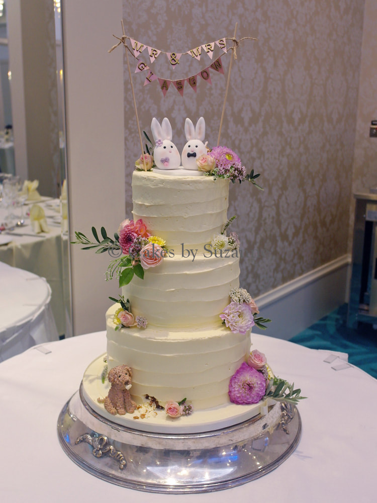 three tier round buttercream wedding cake with bunting, bunny bride and groom, dog eating cake, fresh flowers