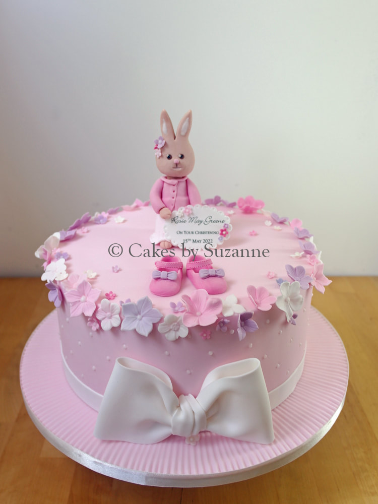 pink christening cake with bunny holding message plaque, baby shoes, blossoms and large bow
